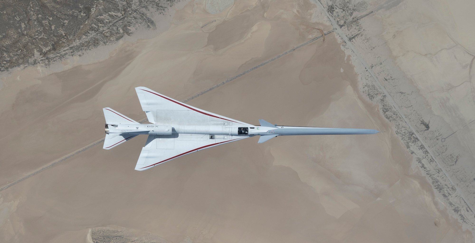 NASA’s white, blue, and red X-59 research aircraft flies over the Mojave Desert near NASA’s Armstrong Flight Research Center in Edwards, California, during its third flight. The aircraft is centered in the frame, viewed from above with the canopy, both wings, NASA and X-59 logos visible, and the landing gear down with one wheel visible. Below, rugged terrain transitions into the smooth texture of a dry lakebed as the aircraft, nearly 100 feet long, flies toward the right side of the frame.